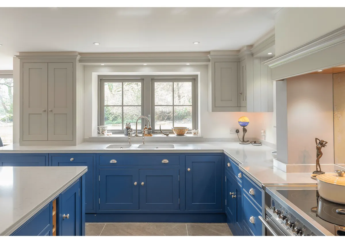 Blue in-frame kitchen under a sash window, with a white worktop and cup handles