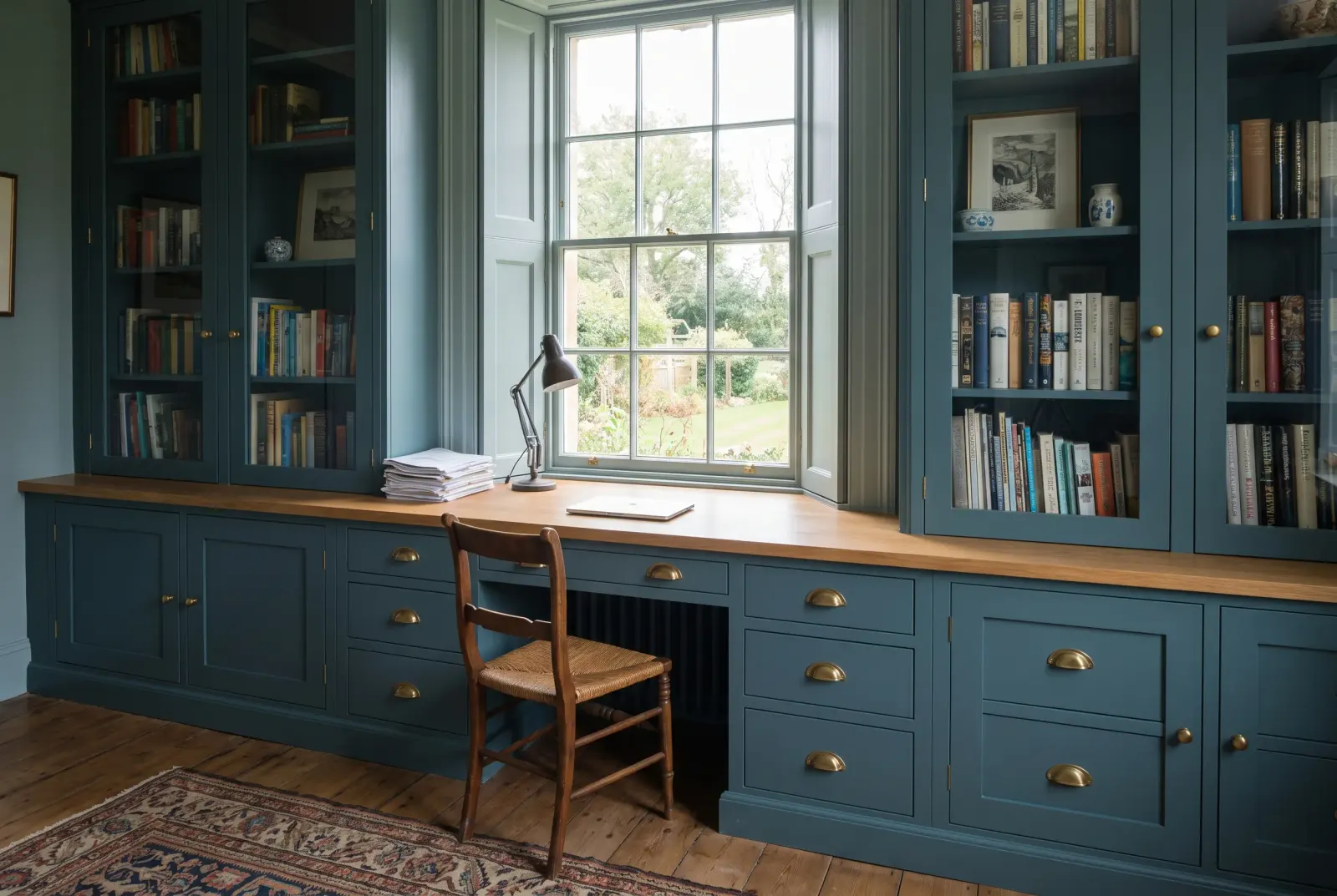 A fitted home study in deep blue-green in-frame cabinets: a desk with an oak worktop built in under a sash window, banks of drawers with brass cup handles, and glazed bookcases either side